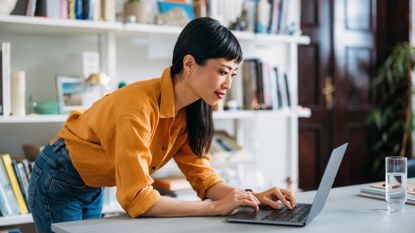 Woman typing on laptop at desk standing up, representing using productivity apps at home