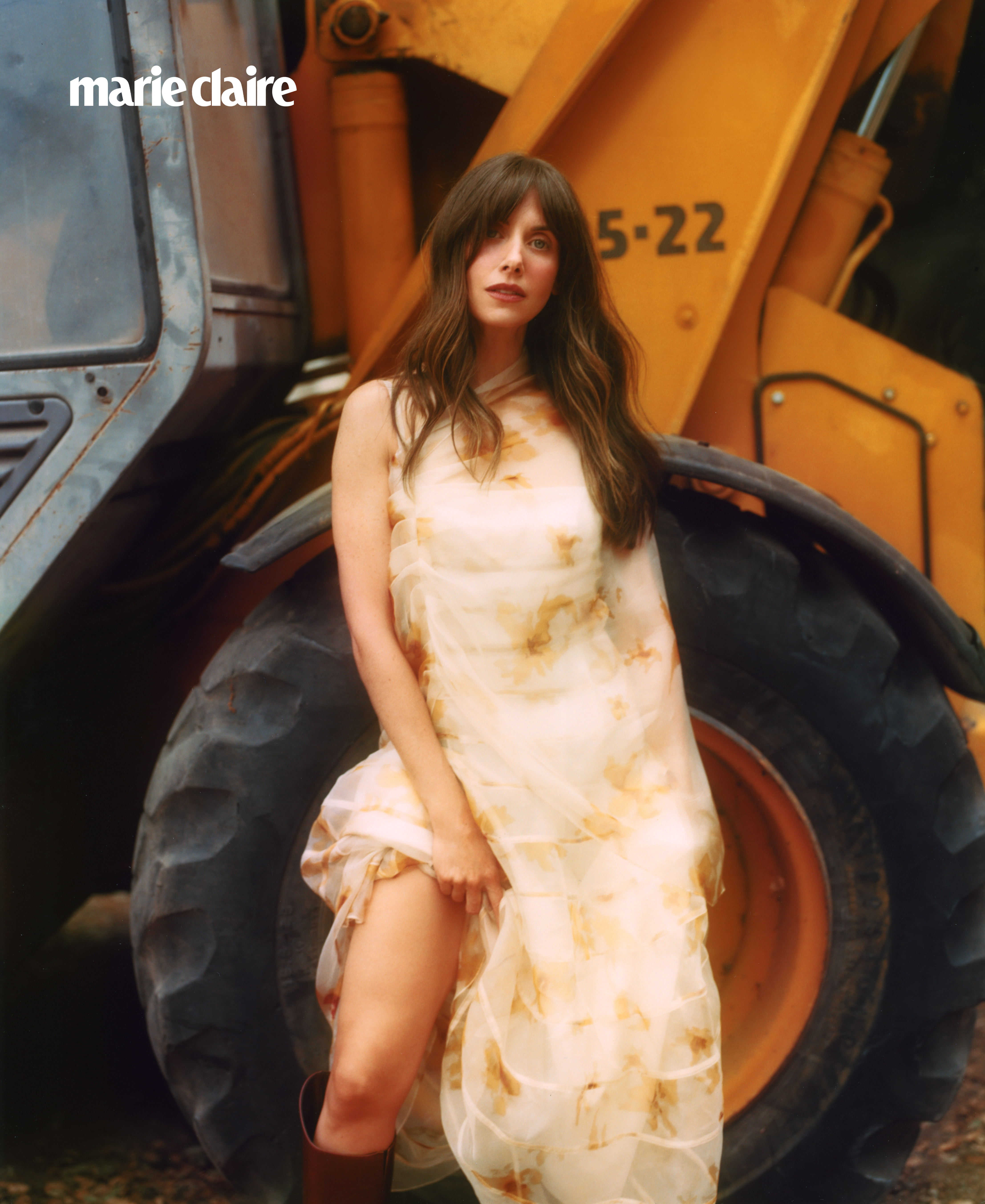 Alison Brie wearing a floaty white dress by a tractor