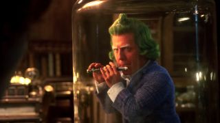 Hugh Grant's Oompa Loompa plays a flute, while held captive under glass, in Wonka.