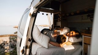 A couple laying in bed in the back of an RV with seafront view