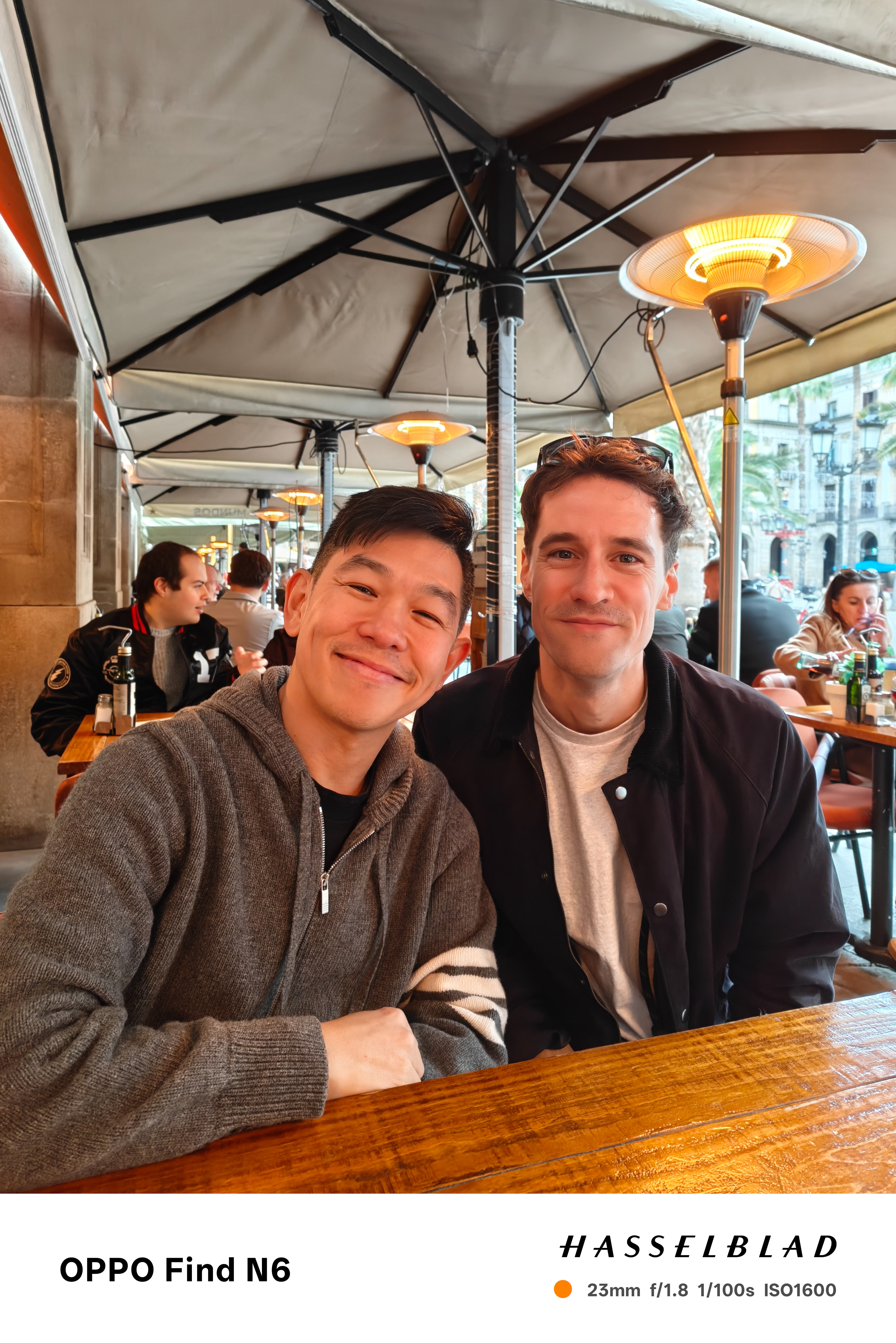 A candid medium shot of two men sitting at a wooden table in an outdoor cafe. They are both smiling at the camera, wearing casual jackets. Overhead, warm heat lamps are visible under the cafe's large tan umbrellas.
