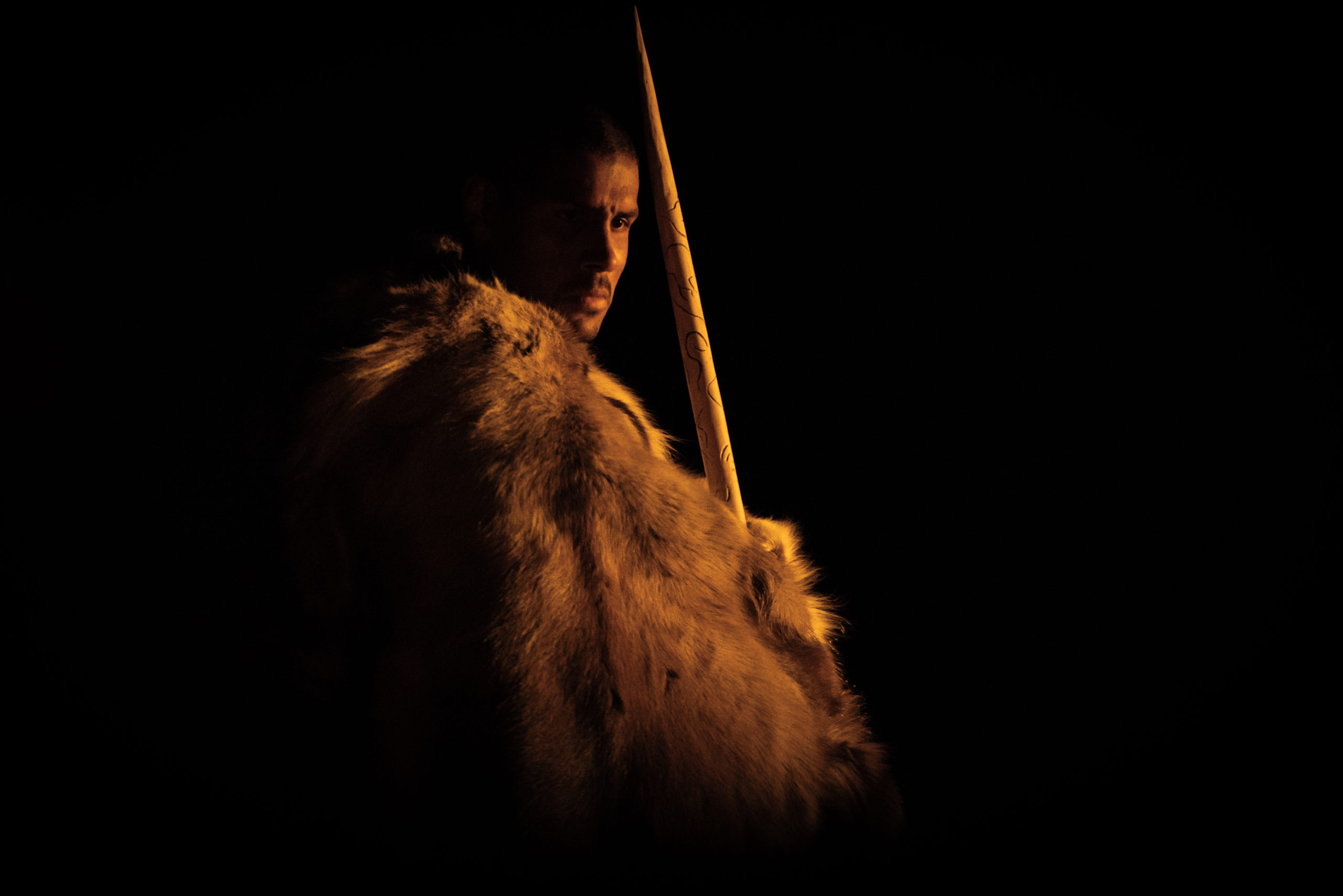 a man wearing a fur jacket holds a long carved stick which ends at a point