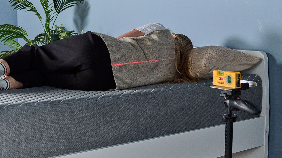 This image shows our sleep tester Nicola Appleton lying on the Siena Memory Foam Mattress during the pressure relief test. We can see her back as she lies on her right hand side on the mattress, using a pillow for support. A laser, balanced on a tripod, shines a laser line on her spine. Her spine is straight, like the line, indicating good spinal support.