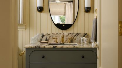 A butter yellow bathroom with wall panelling and a dark green cabinet with marble counters. 