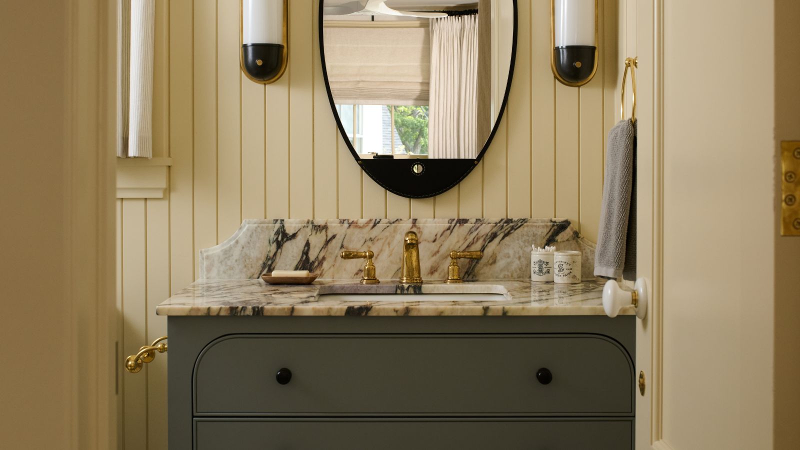 A butter yellow bathroom with wall panelling and a dark green cabinet with marble counters. 