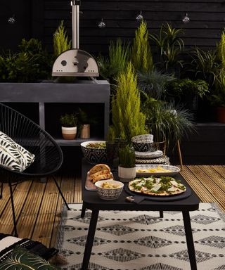 An alfresco dining area with pizza oven, black outdoor dining table, outdoor rug and outdoor plants