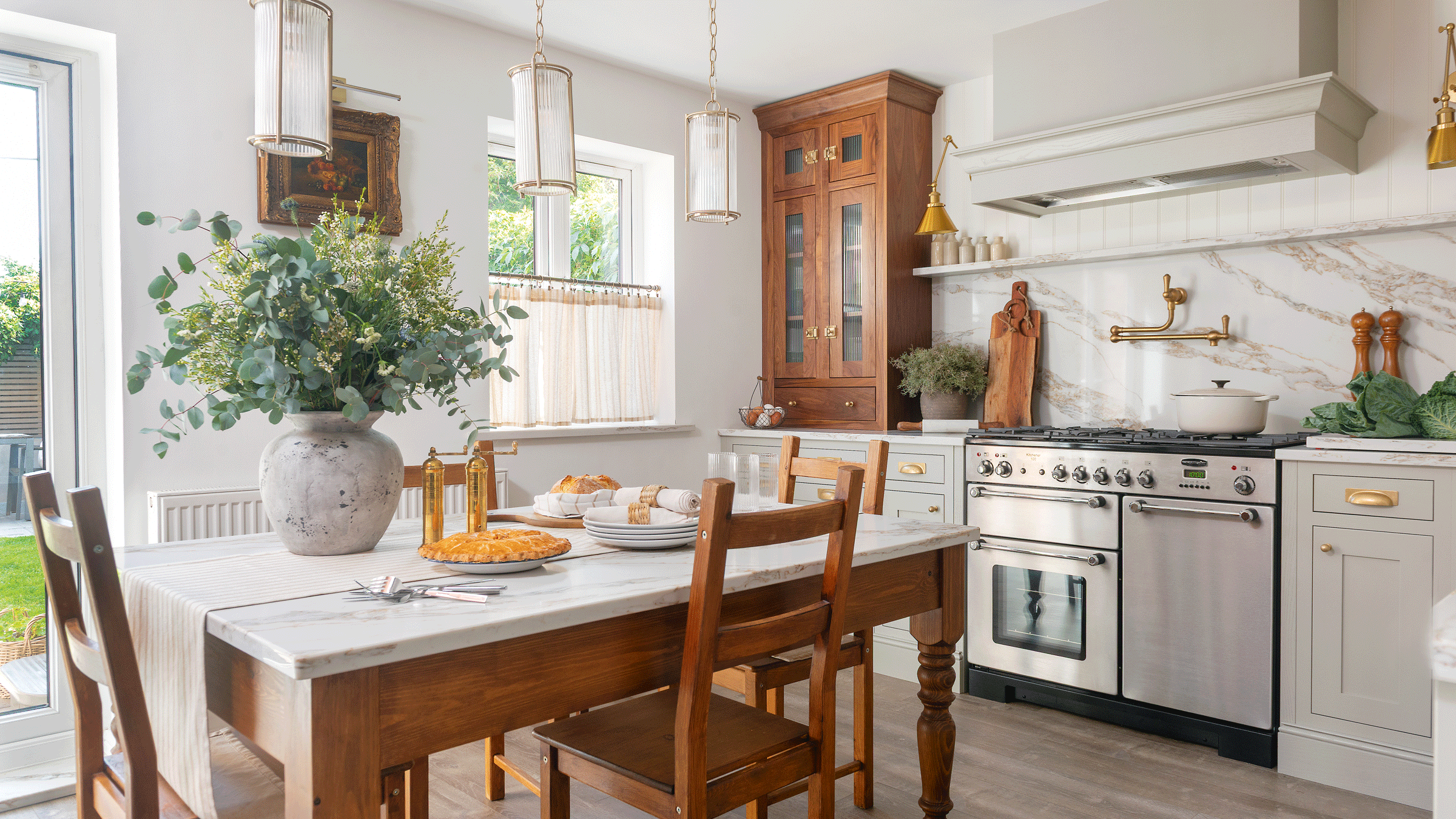 a neutral country-style kitchen with a farmhouse table and chairs in the middle a large range cooker pale green cabinetry and a marble splashback