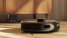 Black iRobot Roomba robot vacuum on wooden floor in front of wooden furniture, with small charging base pictured beneath furniture.