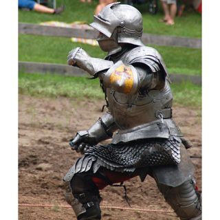 Photo of a person dressed in a knight's suit of armour in a battle enactment, with people sitting on the grass watching in the background