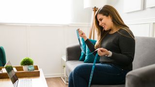 Woman sitting on sofa conducting video call on laptop holding measuring tape