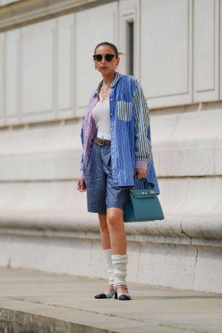 Gabriella Berdugo wears sunglasses, a necklace, a purple and blue striped oversize colored shirt from Ralph Lauren, a white tank top, a brown leather belt, a Hermes blue Kelly bag, navy dark blue striped bermuda / shorts from Cos, knitted mittens ribbed wool socks, blue shoes from Jonak, during a street style fashion photo session, on May 12, 2024 in Paris, France