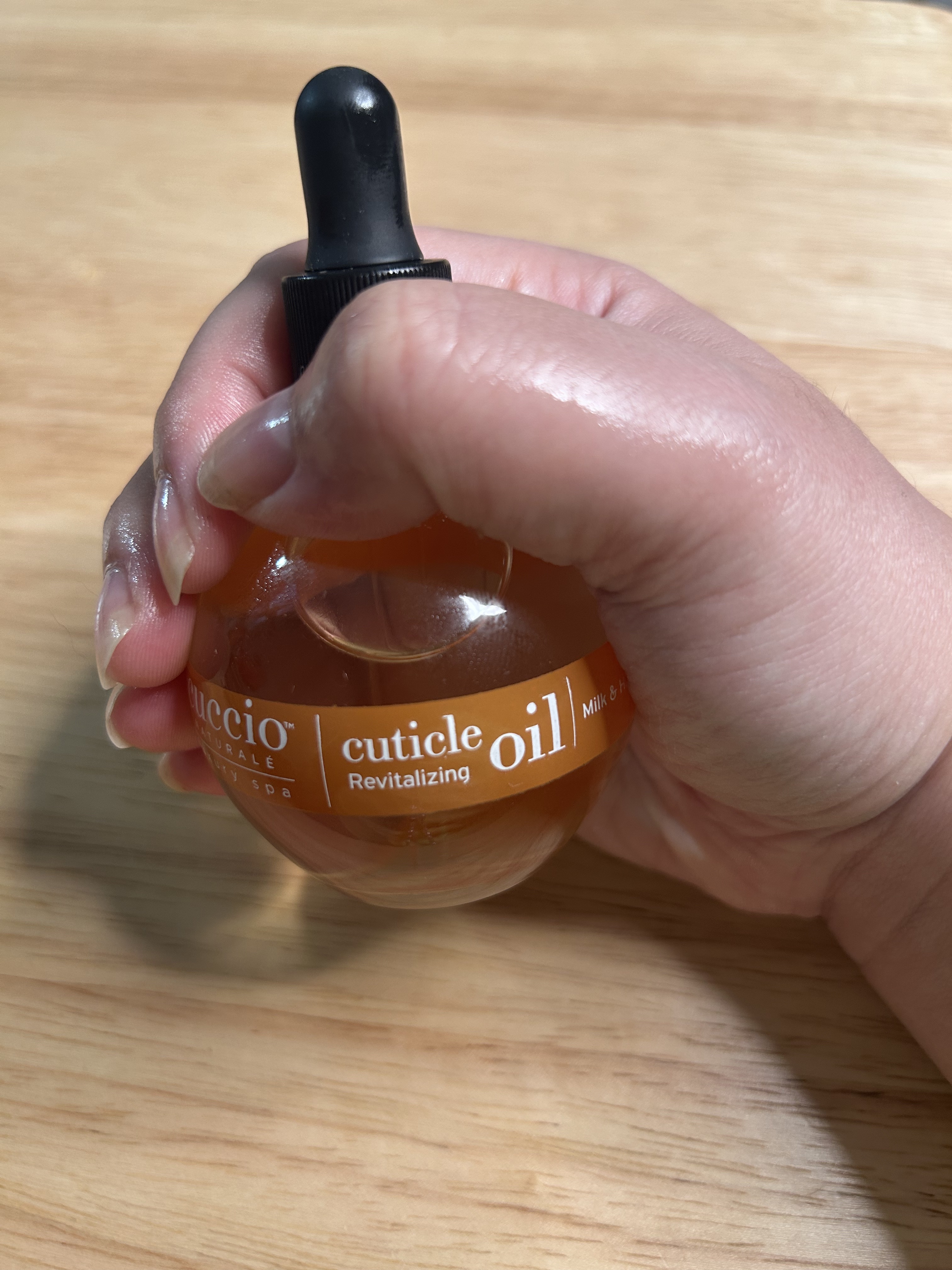 A woman's hand and nails after applying cuticle oil with a wood background holding an orange and black bottle.