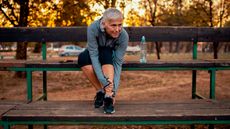 A woman sits on a bench in a park, wearing a running jacket, knee-length leggings and sneakers. She is bending forward and clutching her left shin. Behind her we see the black silhouettes of trees against an orange sunset.