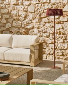 Image of an outdoor seating area that is very minimalistic. The wall is a real stone wall and the furniture is white and light wood. There is a red solar outdoor floor lamp.