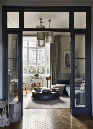 Image of a living room viewed through the glass doorway of another room. There is a blue sectional sofa with a large circular, black coffee table in front of it. There is a circular, gold bar cart underneath the back window and a wooden accent chair beside it. There cylindrical pendant lights hang from the ceiling.