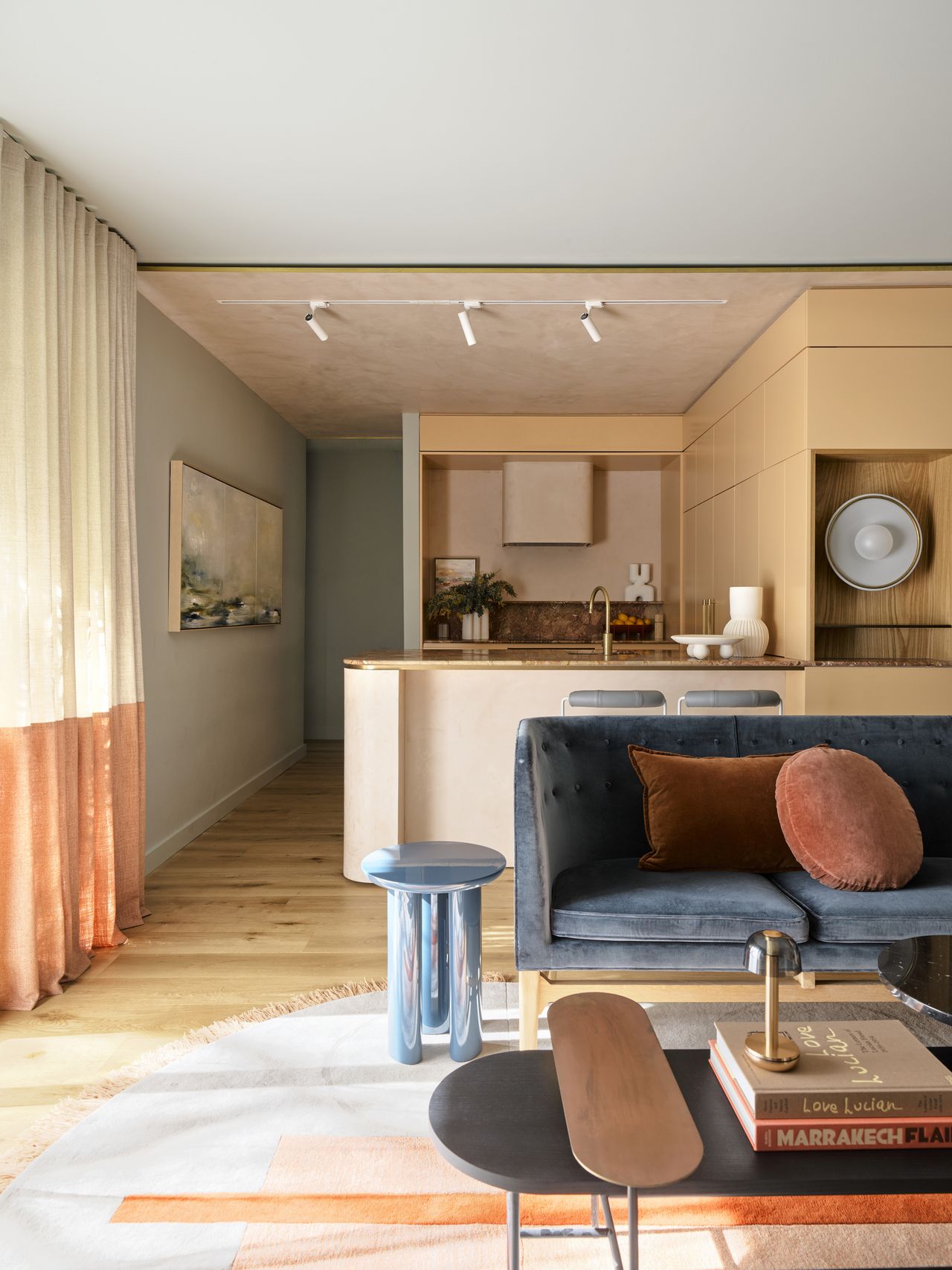 colorful kitchen with peach joinery, textured ceiling, timber floors, blue walls, two-tone curtains, and a living room in front with blue sofa, chrome side table, orange pillows and coffee table styled with books