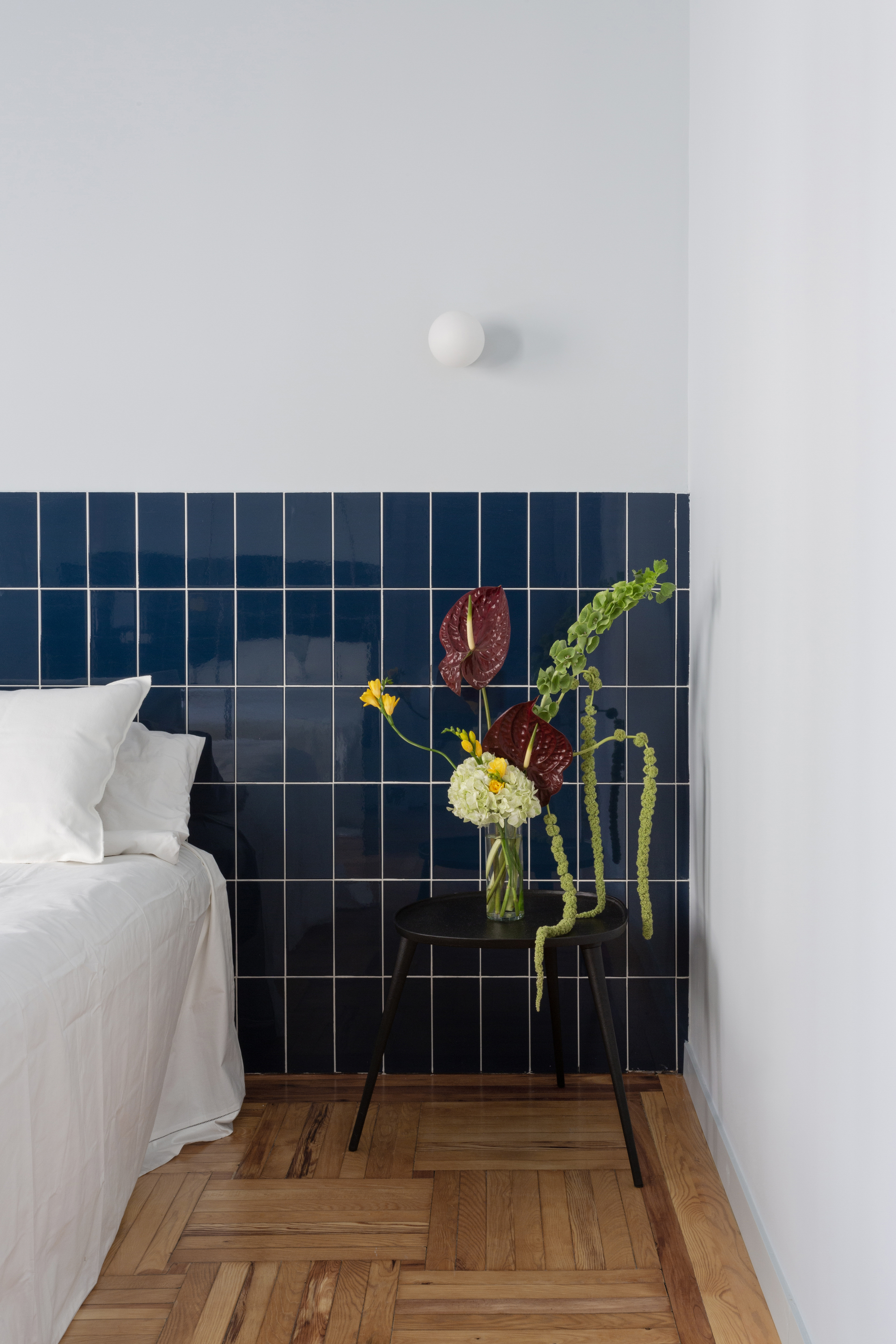 Image of a white bedroom with hardwood flooring. There is a bed with white bedding. The headboard is a navy blue tile that extends the length of the wall. There is a black side table with an artistic floral display.