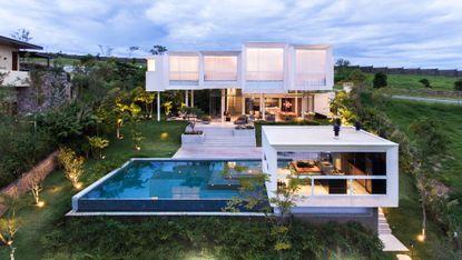 Modern house on multiple levels. A huge pool stands next to one part of the house and floor-to-ceiling windows cover the entire wall. Behind it, the steps lead to the other part of the house, which has the same design, with windows covering the entire wall. 