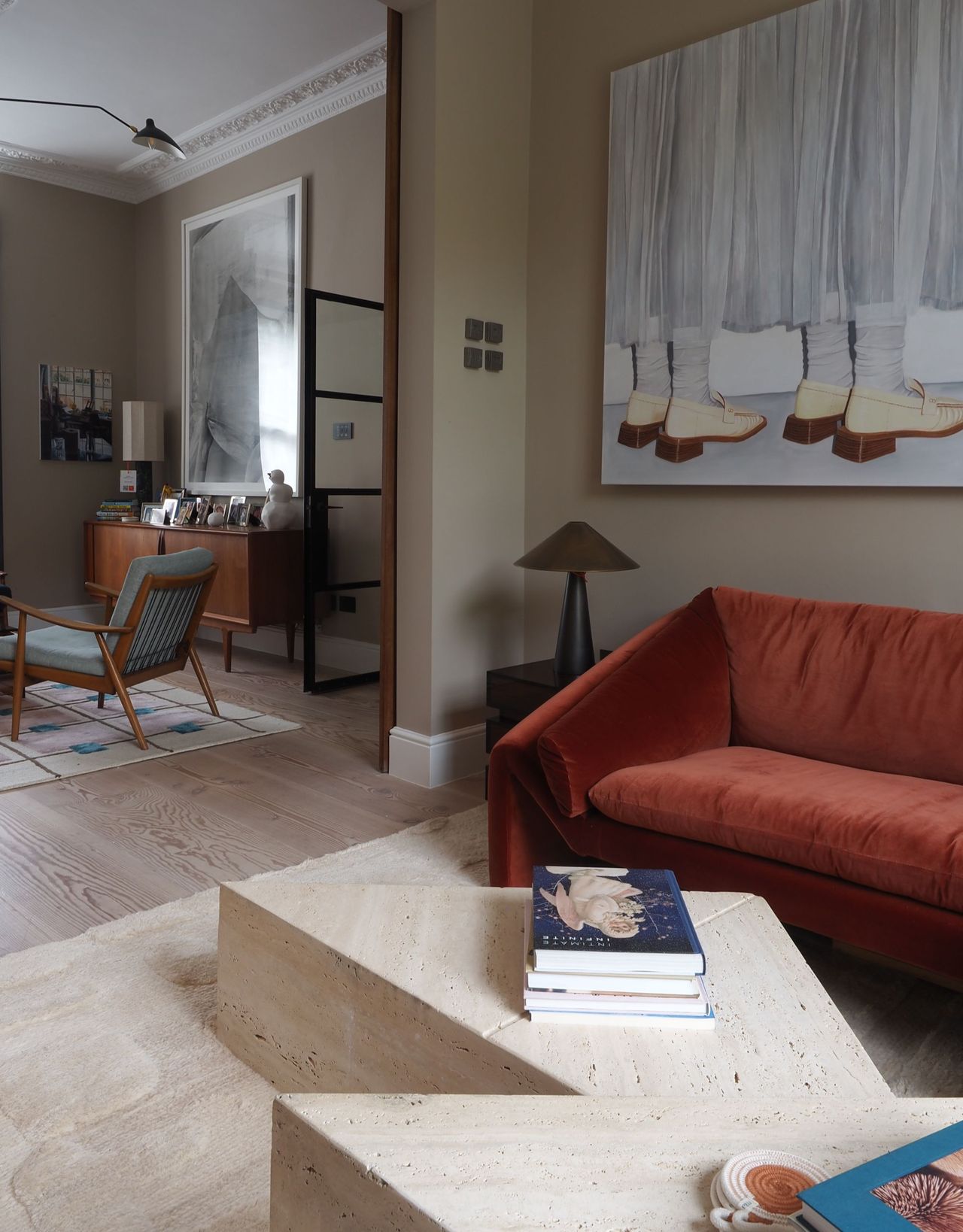 A modern living room with a rust red sofa with an angled armrest, a geometric shaped coffee table with books on top and a large artwork above the sofa with a separate seating area in the background