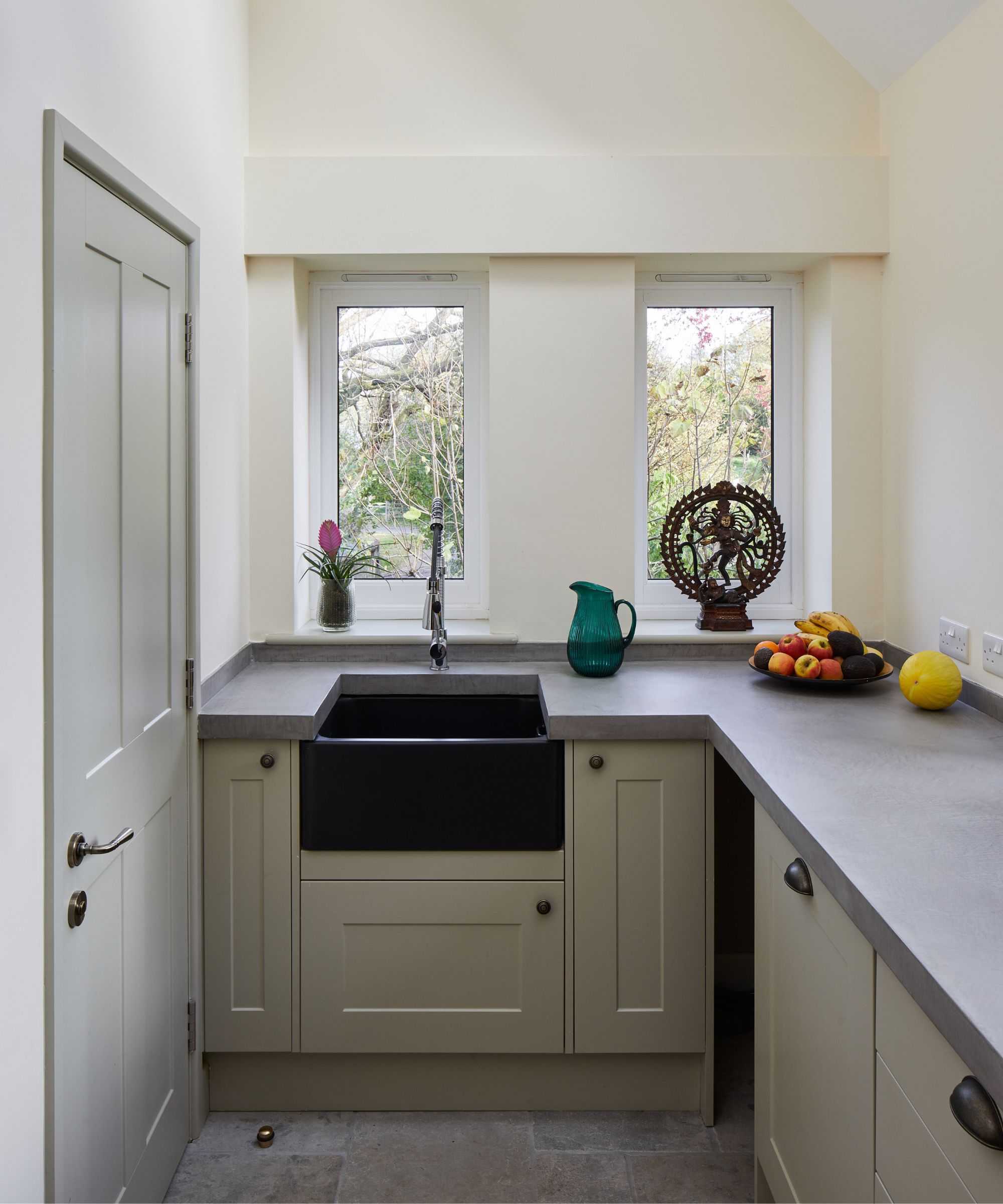 A compact utility room with pale green shaker cabinets, smooth concrete worktops, and a bold black Belfast sink beneath two tall windows. A green glass jug, a fruit bowl, and a decorative bronze sculpture add character.