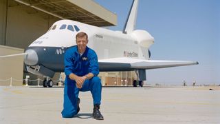 a man in a blue flight suit kneels on a runway in front of a white space shuttle
