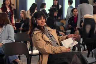 Sasha Bhasin as Praveena wearing a trench coat sitting in a lecture in episode 201 of XO, Kitty.