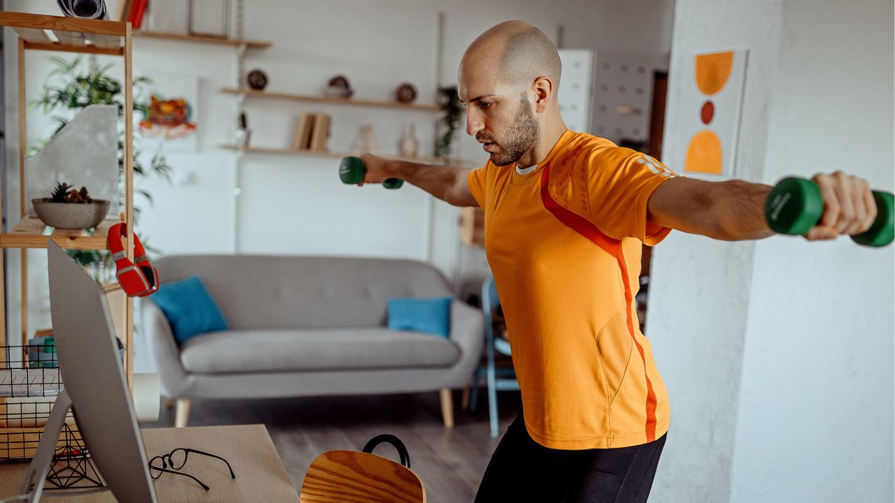 man wearing an orange tshirt holding a light dumbbell in each hand with arms raised laterally at shoulder height. He&#039;s in a living room setting in front of a desktop screen.