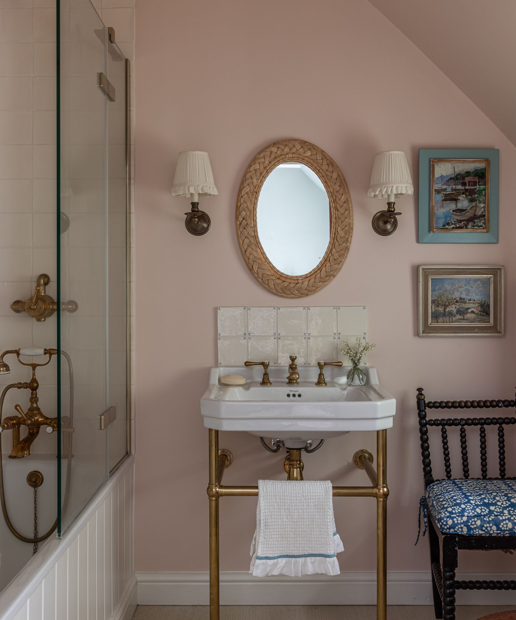 A traditional bathroom with plaster pink walls, a white sink with brass fittings, white backsplash tiles, a white panelled bath with a shower over it, and an oval wall mirror with two sconces.