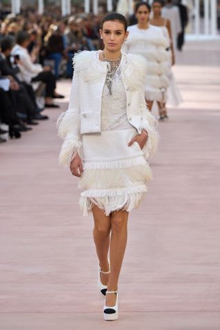 A model at Chanel on the runway during the spring/summer 2025 season wearing a white feather jacket and pencil skirt with platform ankle-strap heels.