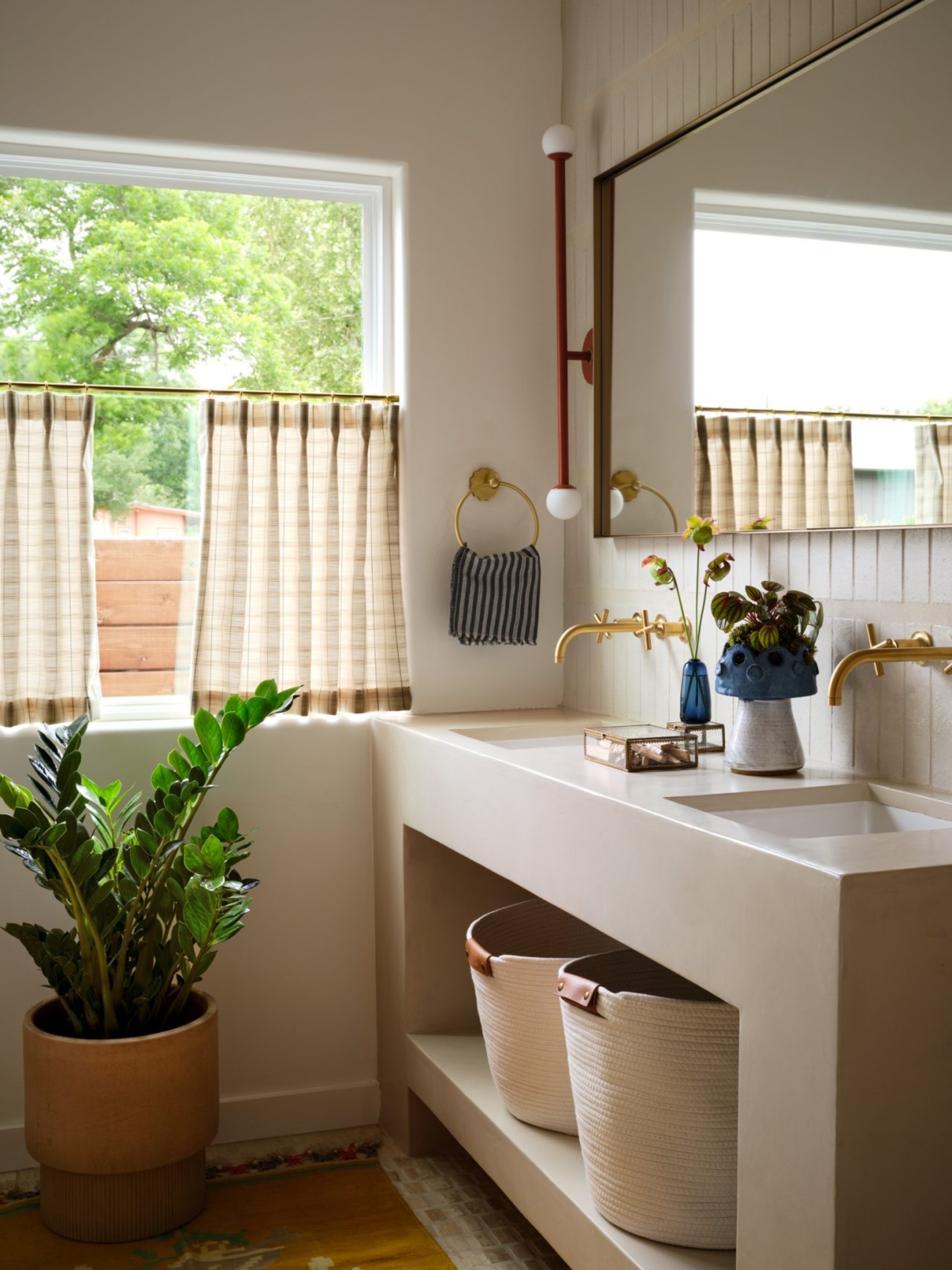 bathroom with built-in vanity with woven basket, wall mirror, gold tapware, wall sconce, cafe curtains over window, plant pot on floor, and vase of flowers