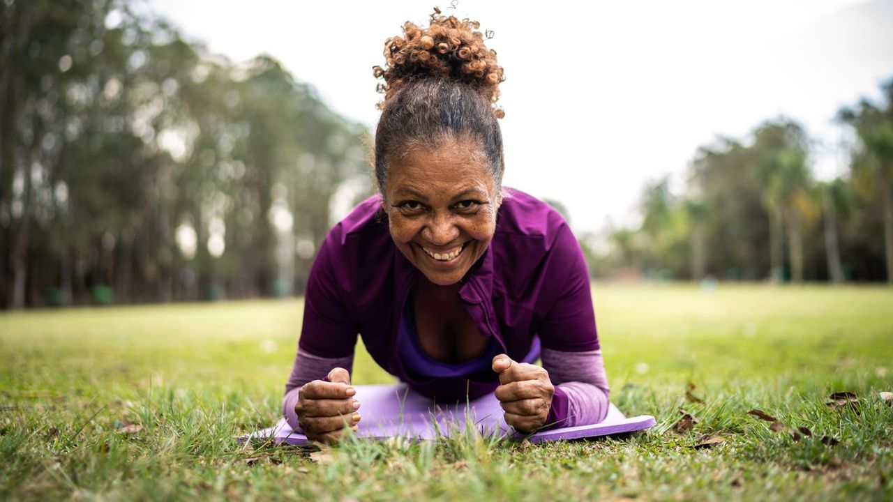 A woman in a park lying on a yoga mat doing a plank and smiling at camera