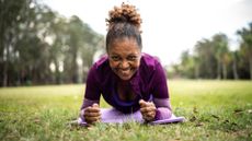 A woman in a park lying on a yoga mat doing a plank and smiling at camera