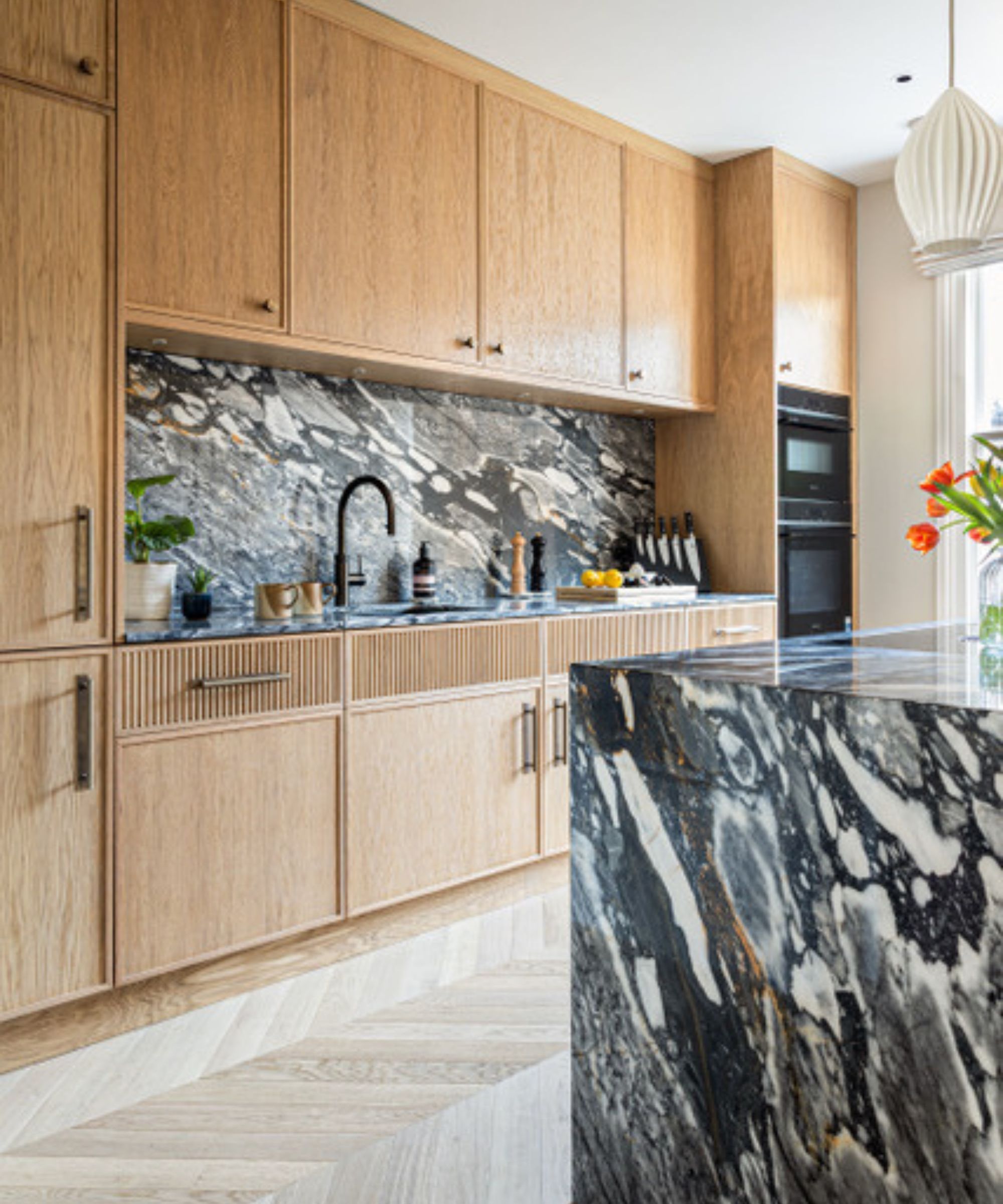A light oak kitchen with dramatic dark grey and white marbled stone worktops and matching full-height splashback, fluted drawer fronts, black tap and oak herringbone floor.