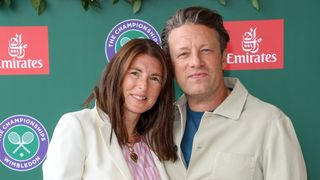 Jools Oliver and Jamie Oliver attend Wimbledon in cream jackets standing in front of a branding board with their heads together.