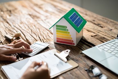 Small model house with energy efficiency grades printed on the side, while a person is seen writing on a clipboard and using a calculator