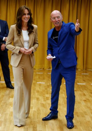 Kate Middleton wearing a beige trouser suit standing on a brown floor next to a man in a blue suit who is pointing ahead