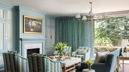 Blue-green Colorado family room with large windows overlooking a lush landscape, painted molding, geometrically patterned club chairs, and a multi-shade chandelier overhead