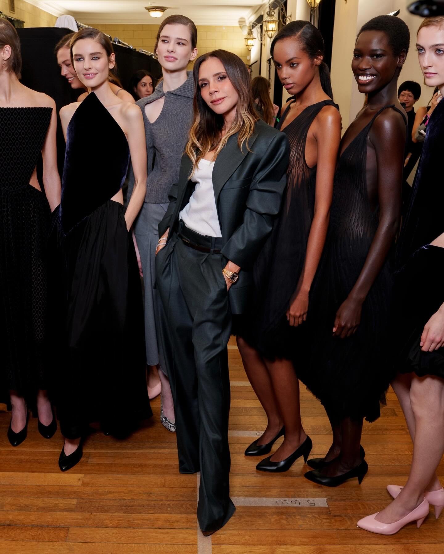 Victoria Beckham poses with models after the Victoria Beckham AW26 runway show wearing dark grey puddle pants with a grey blazer and a white tank top.