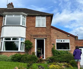 The exterior of a red brick semi-detached house with bay windows, mature front garden shrubs and a person walking towards the open front door.