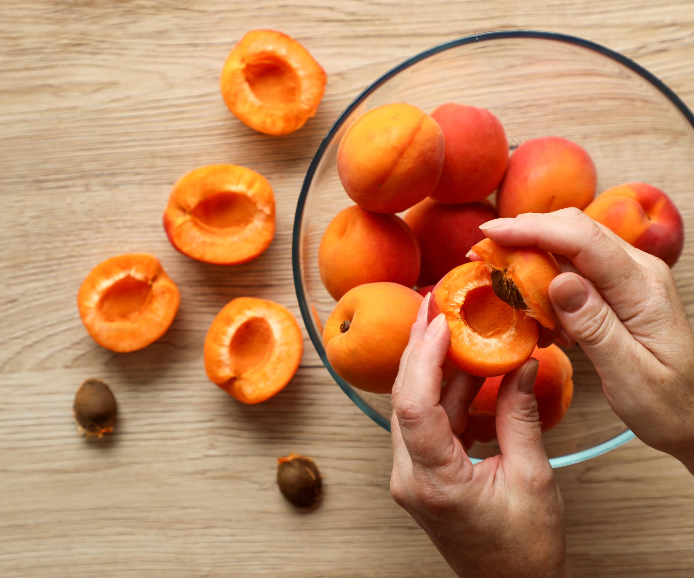 Hands splitting open one of several apricots over a glass bowl