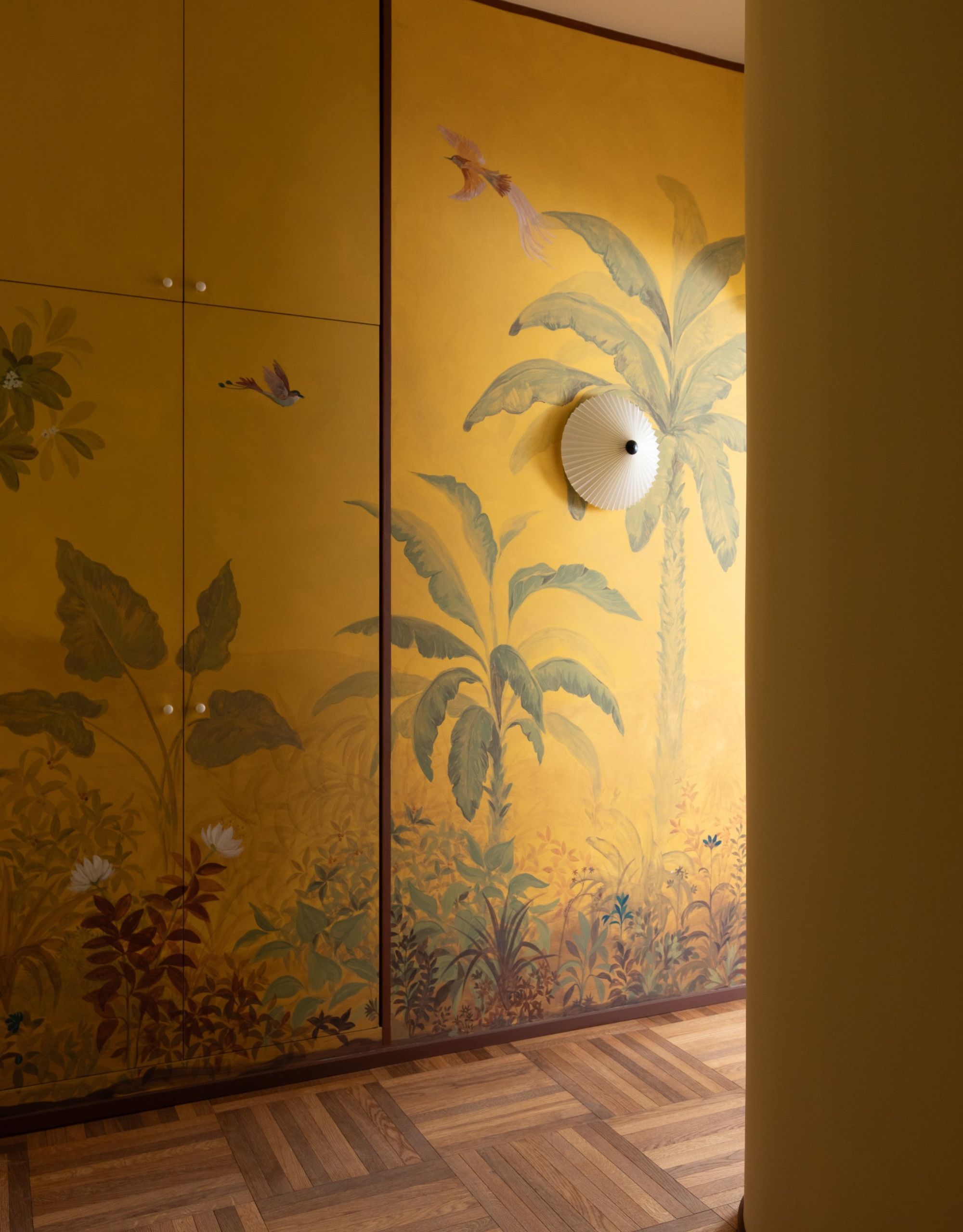 A yellow-painted hallway mural conceals cubpoards, and there's a white pleated wall light on the painted wall next to the cupboards over the top of the mural