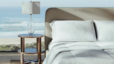 A Saatva bed made up with white bedding beside a wooden nightstand with a glass lamp and a white lampshade. Behind is a beach landscape.