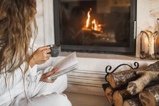 A woman holding a cup of coffee while reading a book in front of a cosy fireplace.