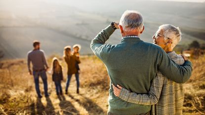 An older couple with their arms around each other look toward their family in a field.