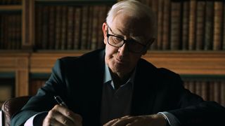 John le Carré (David Cornwell) sits at a desk wearing glasses while writing something down with books behind him