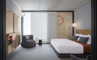 A room in the Nobu Hotel Shoreditch. The room is decorated in various shades of gray - walls, carpet, and bedside tables are in lighter gray, while the armchair and a small coffee table are in darker gray. The headboard is painted gold, as well as the mini bar across from the bed. Floor-to-ceiling windows cover the far wall.