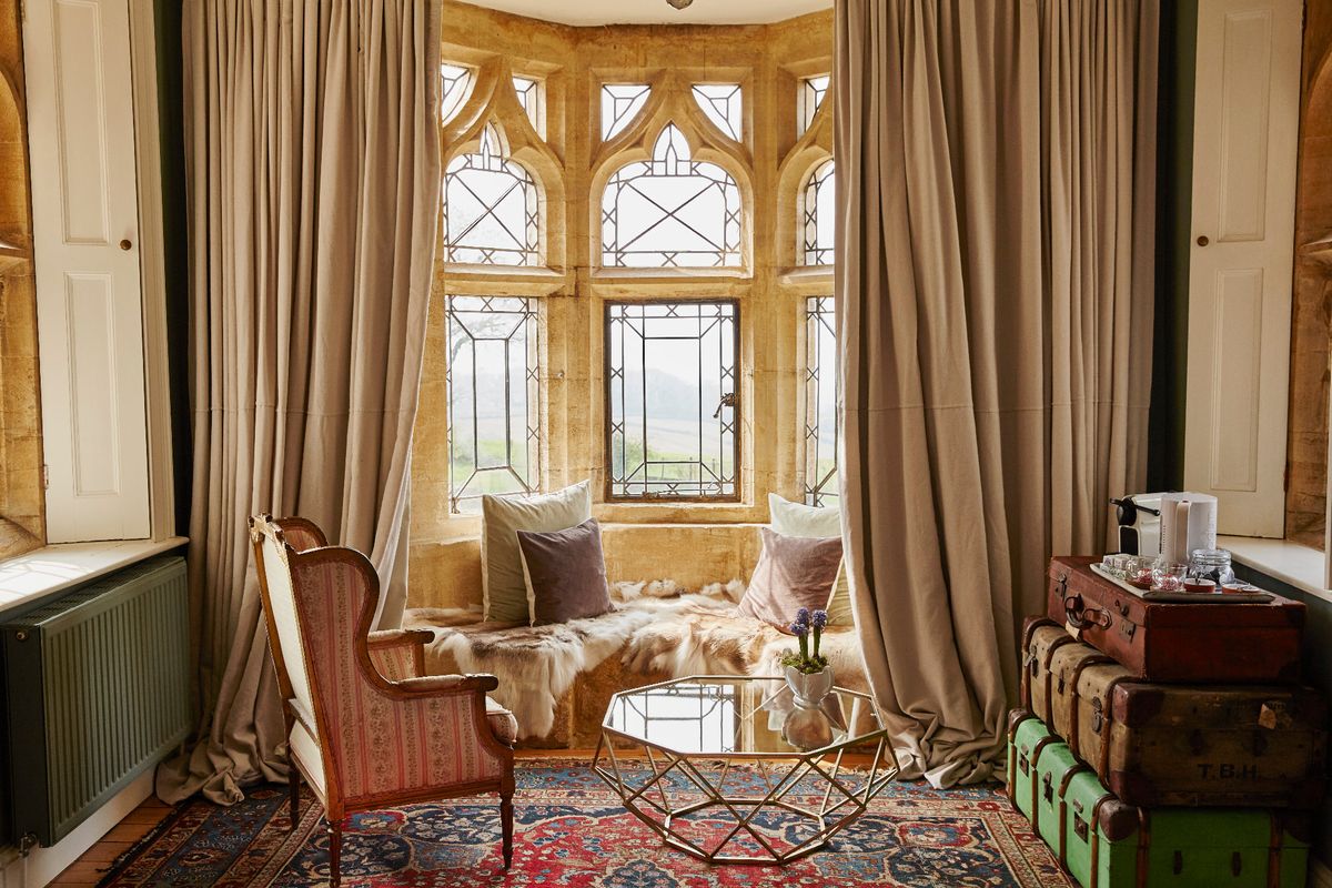 Seating area with wing armchair, colourful rug, piled luggage trunks and a fur covered window seat by a gothic stone and leaded window.