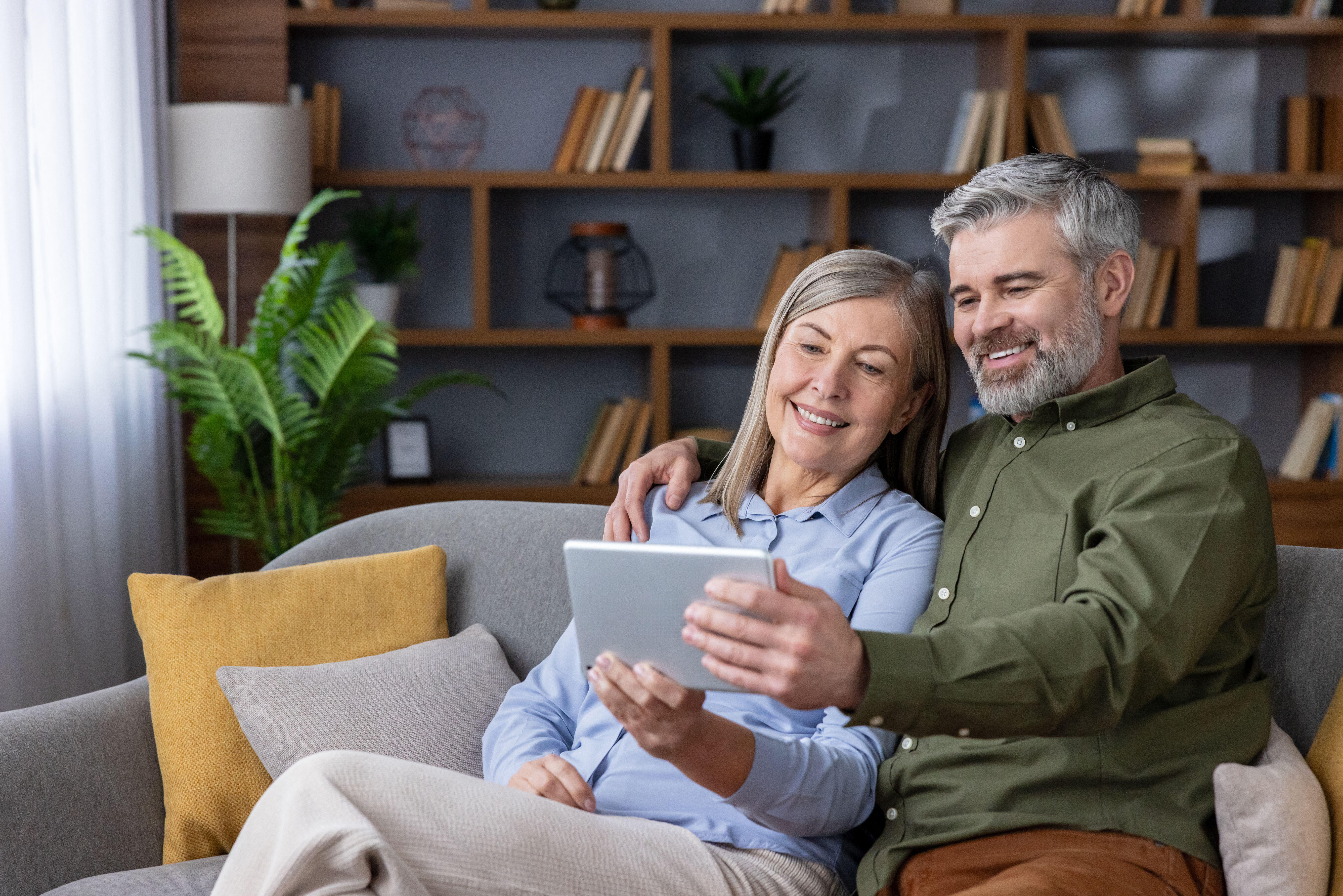 3E3R3B4 Happy senior couple relaxing on a comfortable sofa in their modern living room, smiling while browsing content on a digital tablet together, connectin