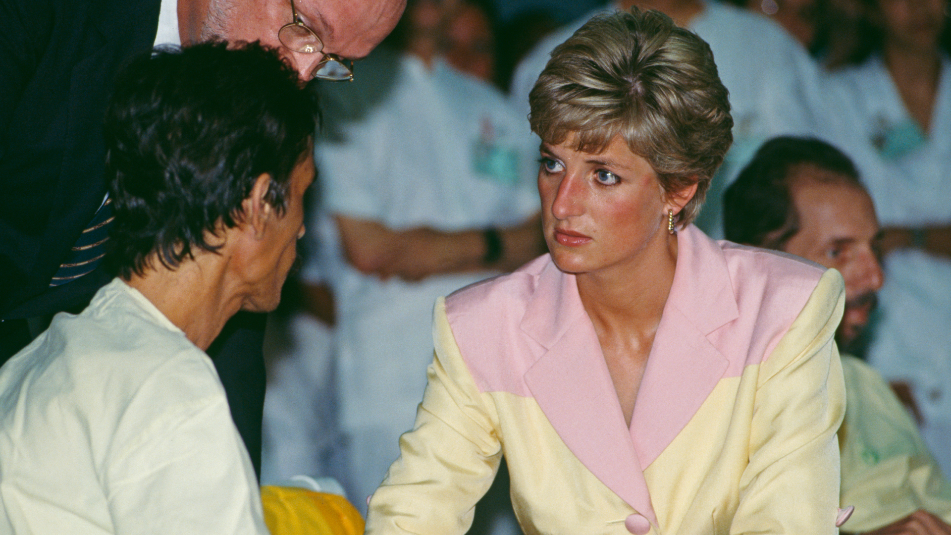 Princess Diana in a yellow and pink blazer talking to a man in a hospital gown