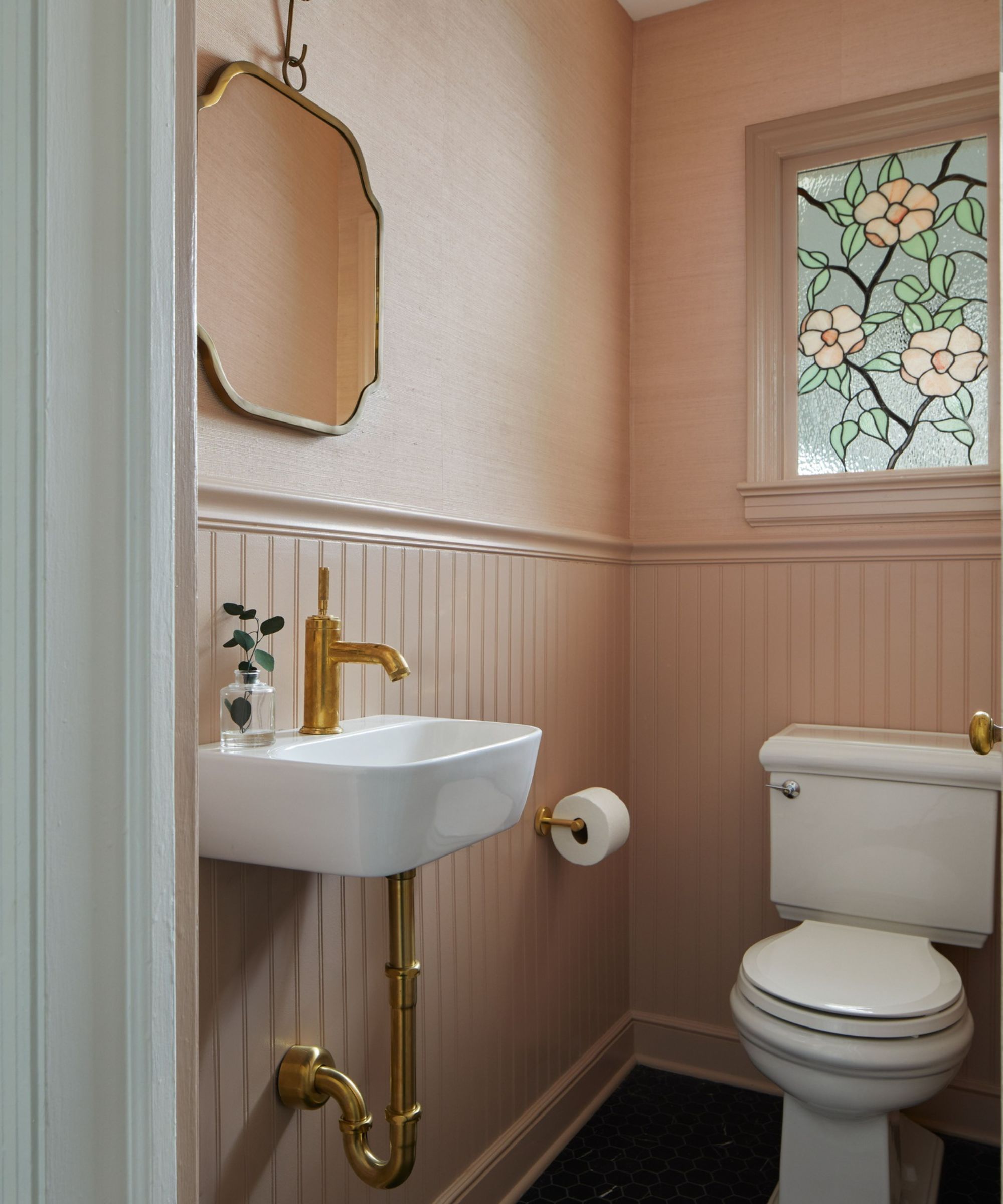 A powder room with plaster pink panelling, with a matching grasscloth wallpaper on the upper walls and a stained glass window.